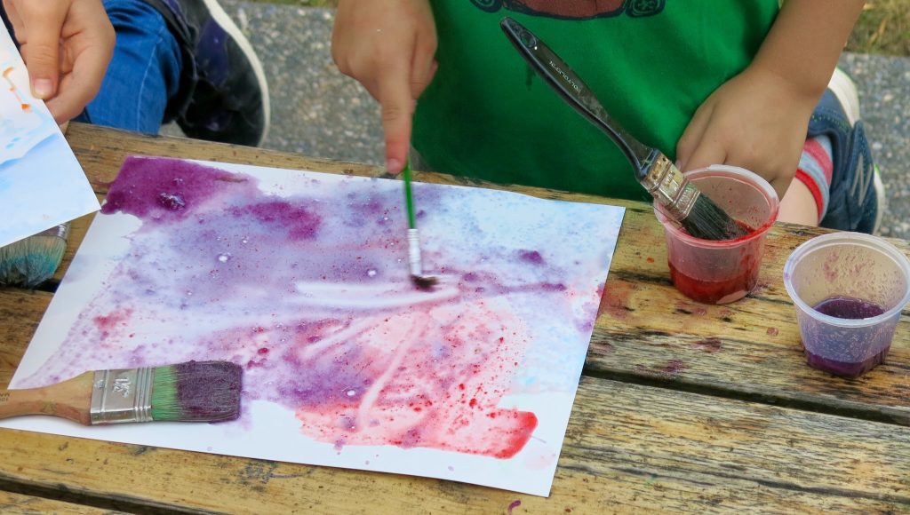 A child holding a paintbrush, with small containers of colored liquid to one side.