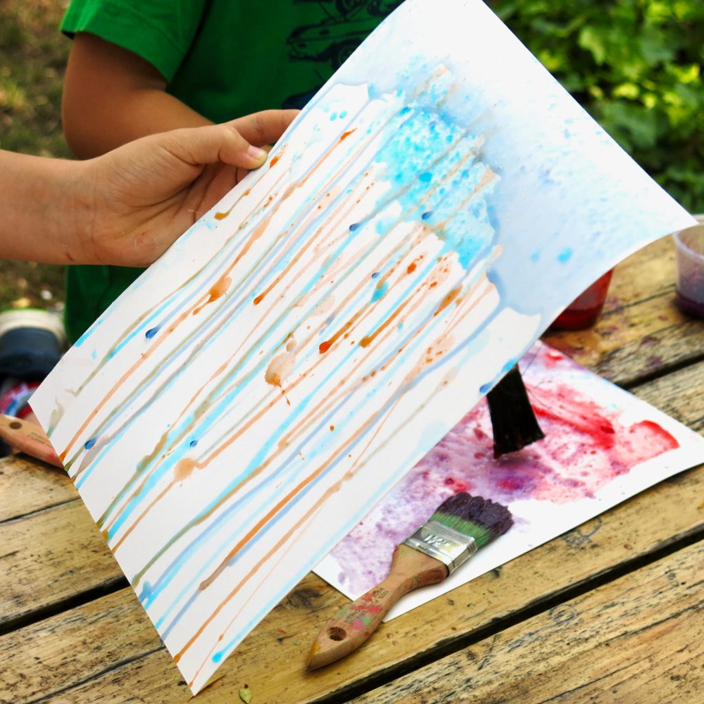 A hand holding a stiff sheet of paper on which colored liquid is dribbling down.